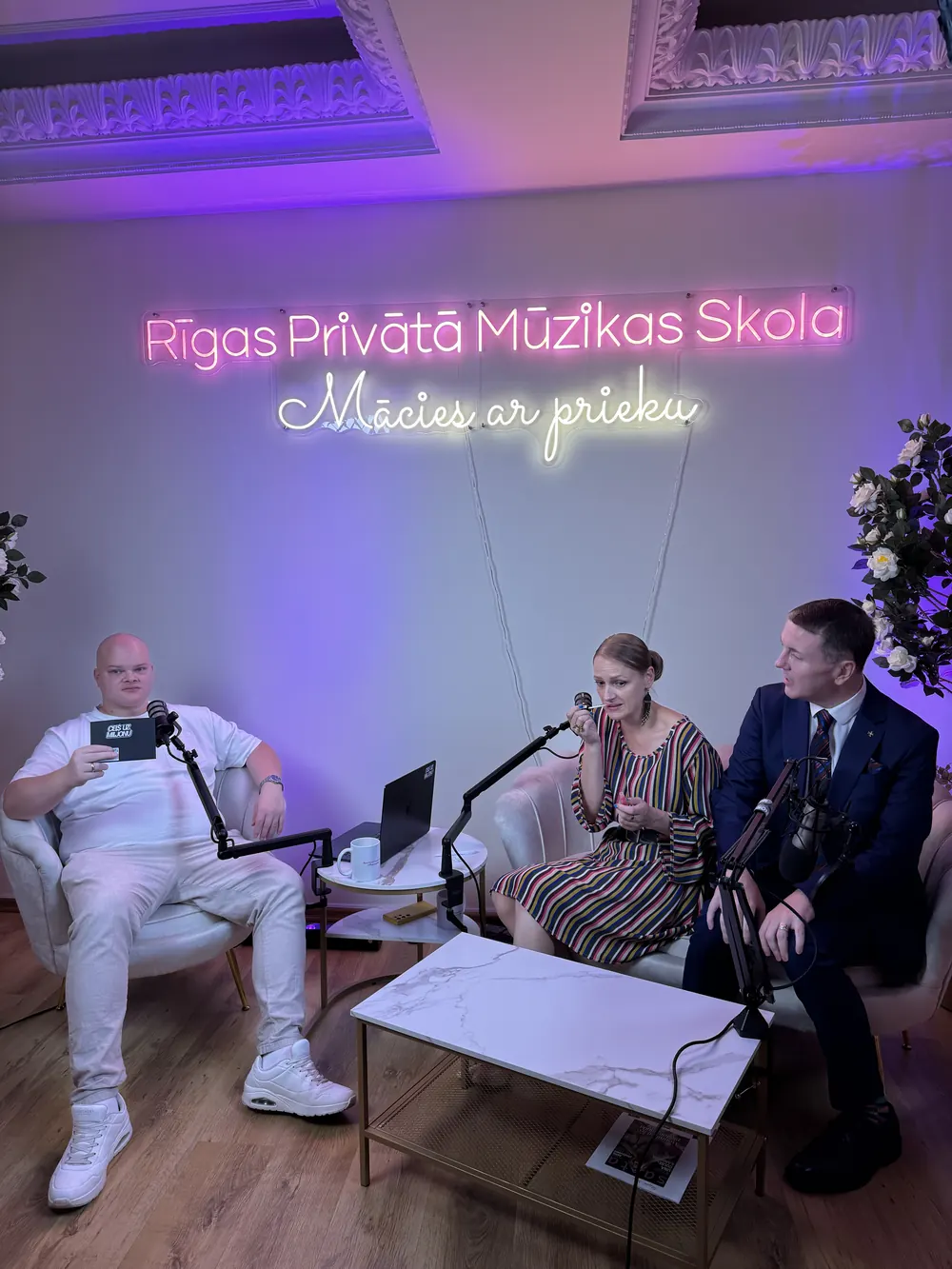 Rīgas Privātā Mūzikas Skola founders being interviewed on camera in the studio, beneath the school's neon sign
